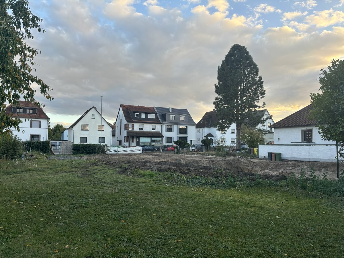 Unbebautes Grundstück mit Wiese und ausgehobenem Erdreich, dahinter Wohnhäuser